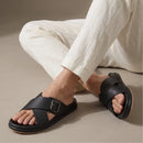 A person wearing black  slide sandals with wide crisscross straps and a single square buckle, seated on a beige floor in white pants.